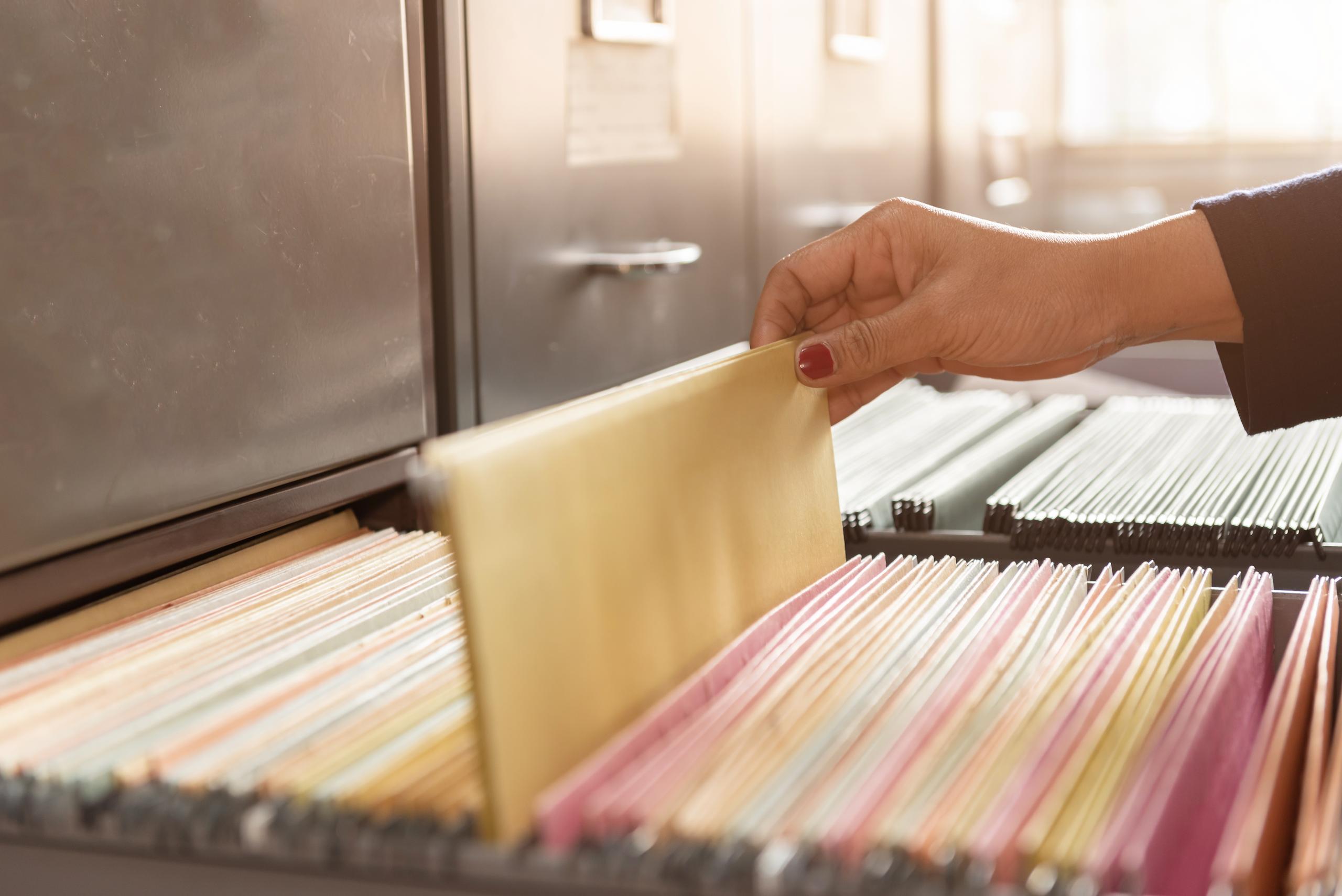 A row of filing cabinets has multiple drawers opened. The drawers contain organized files. A woman's hand is seen pulling a file out of the drawer.