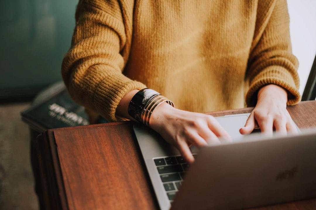 A torso of unknown gender is shown wearing a yellow sweater and bracelets. They are typing on a laptop that is on a wooden desk.