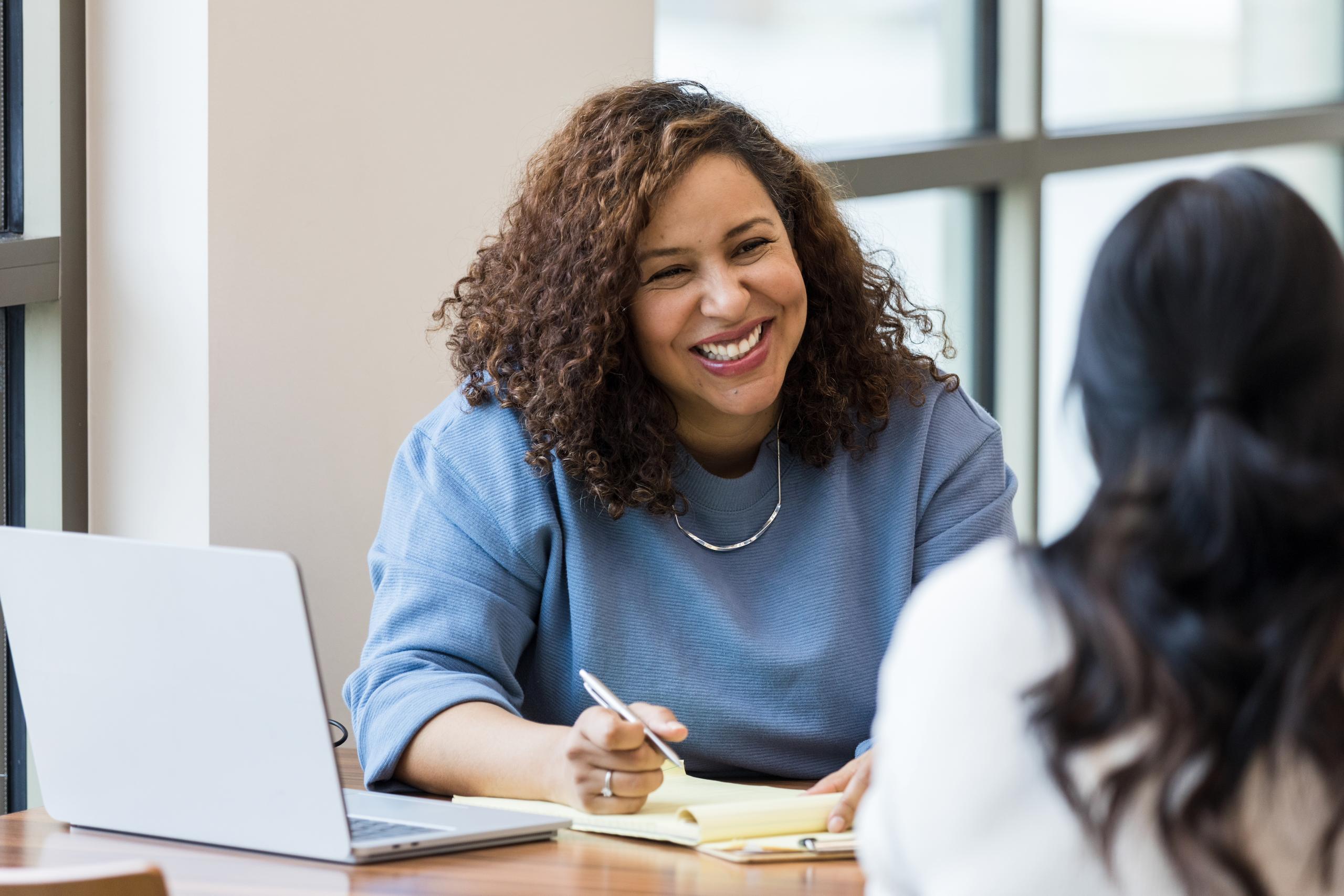 A woman with brown curly hair and a blue sweater sits at a desk smiling with a pen in hand. She is facing another woman with long black hair.